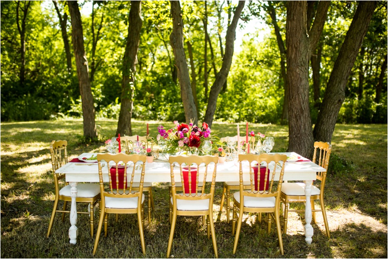 Large wooden table filled with beautiful bouquets, candles, place settings, golden silverware, golden chairs in the forrest