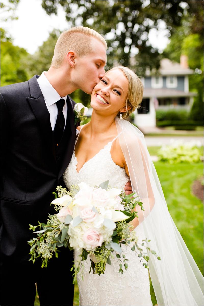 The groom is kissing the bride while she smiles and looks at the camera