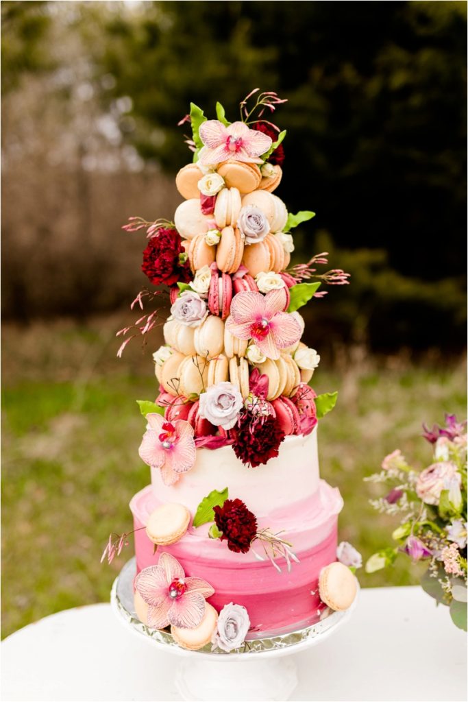  A wedding cake decorated in an ombré pink design, topped with a tower of macarons and adorned with pink orchids, burgundy peonies, and pale roses. The cake sits on a white cake stand.