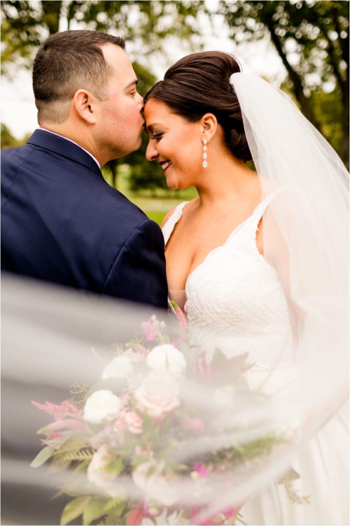 Intimate close-up of groom kissing bride's temple while she smiles, veil and bouquet framing bottom of frame