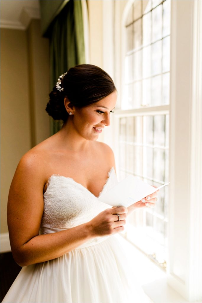 A bride in a strapless white gown stands near a window, smiling as she reads a letter. Soft natural light illuminates her face and dress.