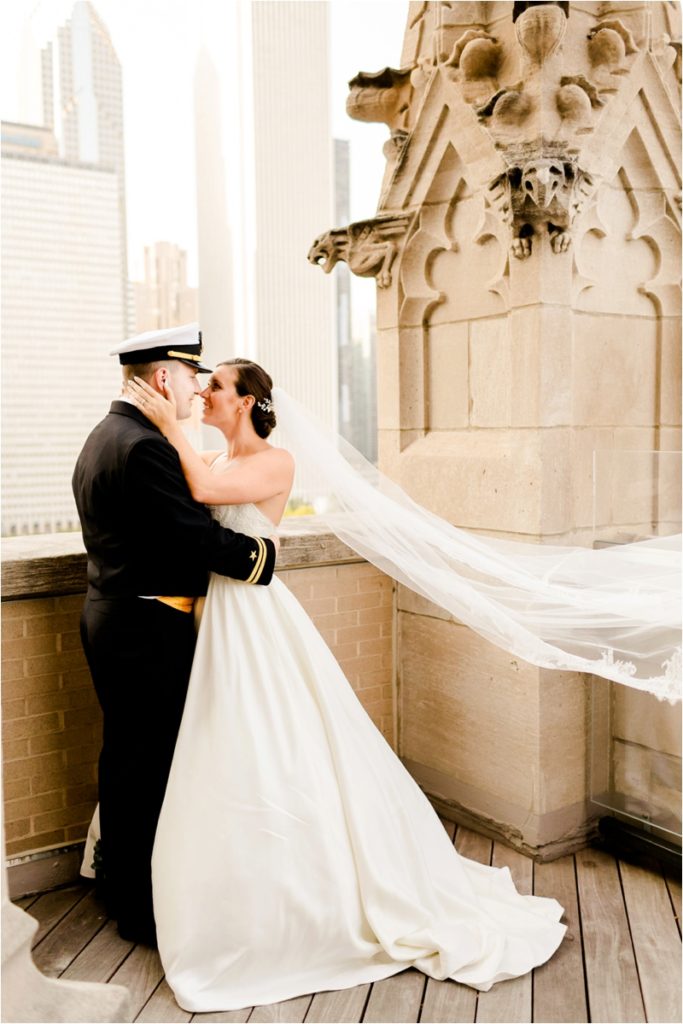 Military wedding pose with bride's veil flowing on balcony, showing natural interaction between bride and groom in formal attire