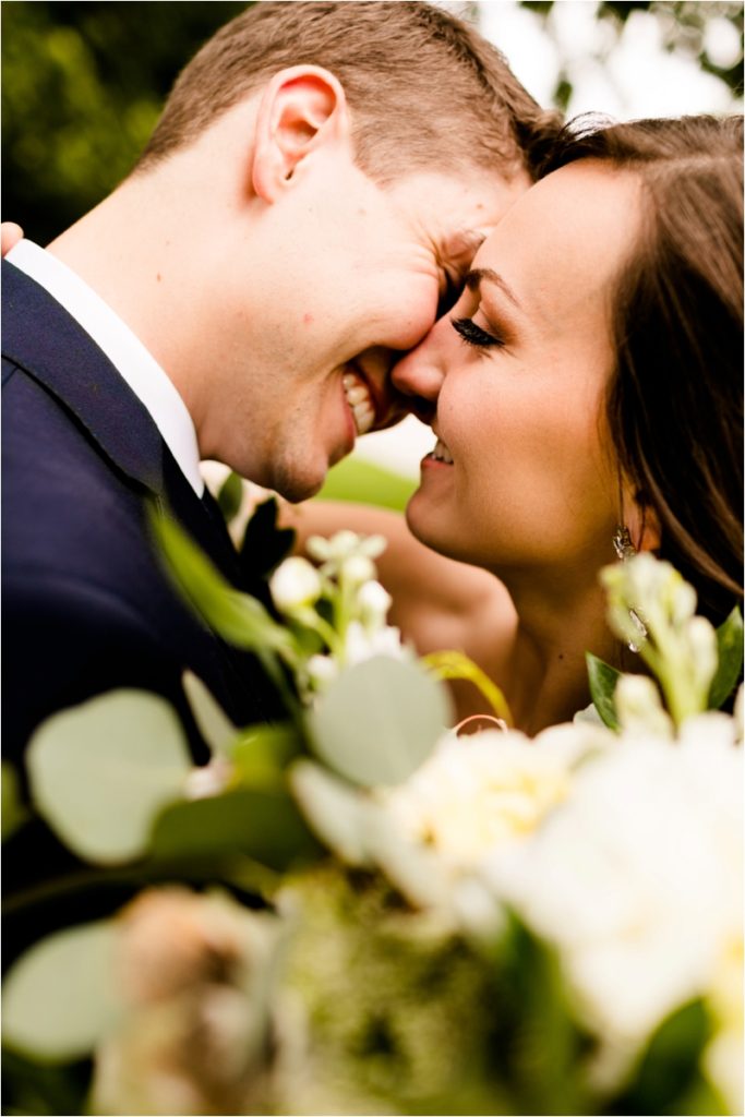 Close up wedding portrait of bride and groom touching foreheads and laughing, demonstrating natural couple poses with white flowers in background