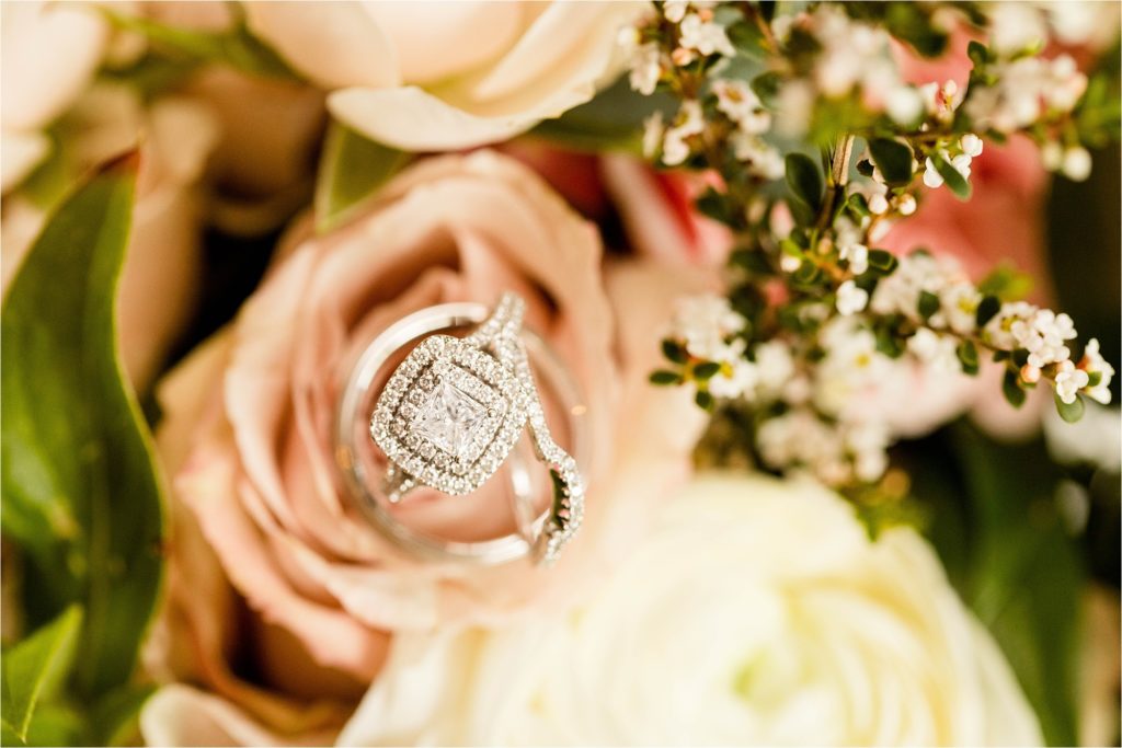 Close-up of diamond halo engagement ring nestled in blush pink rose with white flowers in background