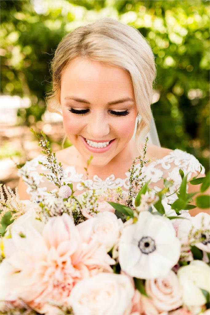 Close-up portrait of smiling bride with blonde hair and lace dress holding bouquet of blush dahlias, white anemones, and delicate greenery