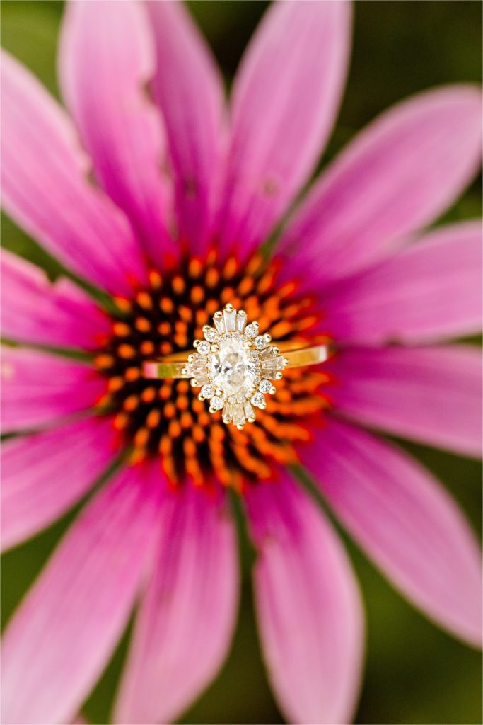 A close-up photograph of an engagement ring with a vintage-inspired diamond setting, artistically placed in the center of a bright pink coneflower bloom.