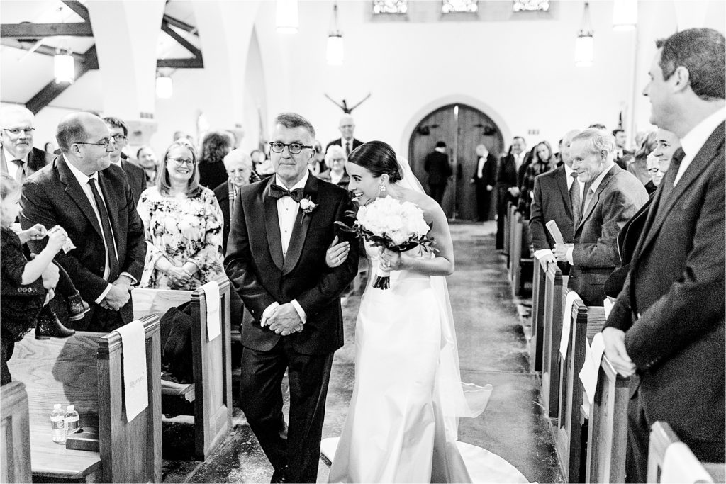  A bride in a white gown walks down the aisle of a church, holding her father’s arm while smiling and leaning toward him. The father, wearing a tuxedo, looks ahead solemnly. Wedding guests on both sides watch with smiles.