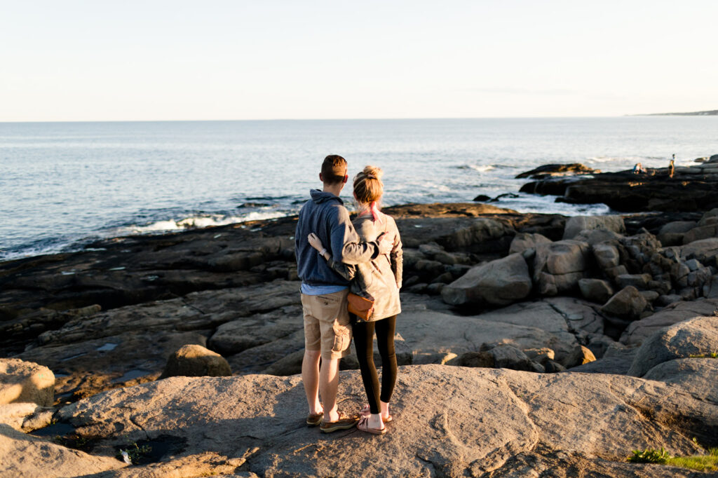 Engaged couple embracing while watching sunset on rocky coastal shoreline during golden hour engagement session