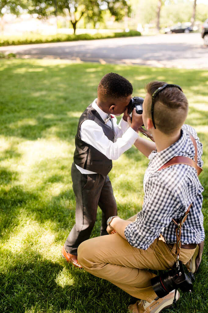 Professional wedding photographer teaching young ring bearer how to use camera during outdoor wedding ceremony