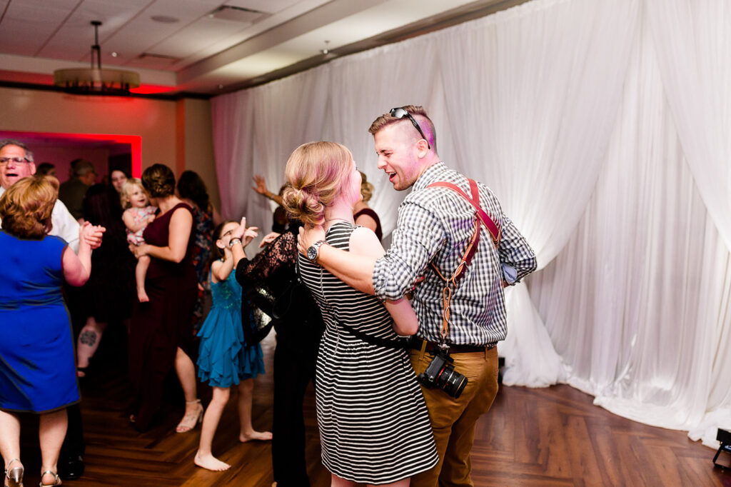 Wedding photographer dancing with second shooter at reception while wearing camera gear and lighting equipment