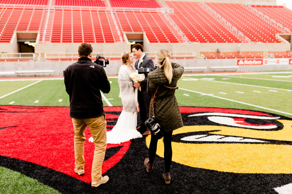 Behind the scenes of wedding photographers capturing bride and groom portraits at a stadium football field with red seats and team logo