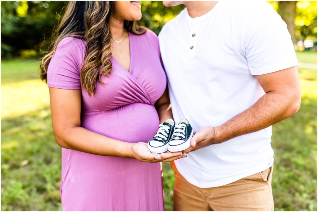 parents show off baby sneakers during maternity photos in Ewing Park photographed by Bloomington IL maternity photographers Caitlin and Luke Photography