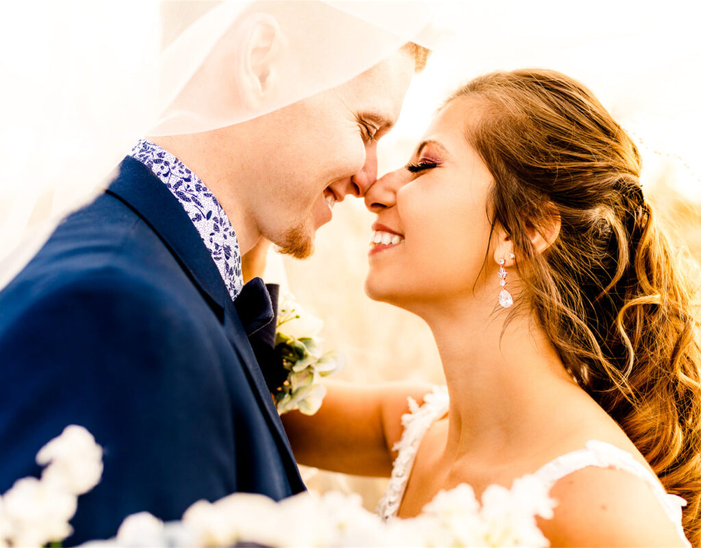  A close-up portrait of a bride and groom about to kiss, backlit by warm golden hour sunlight. The groom wears a navy suit with a patterned shirt, and the bride wears a white dress with dangling earrings. Their faces are close together and both are smiling intimately.