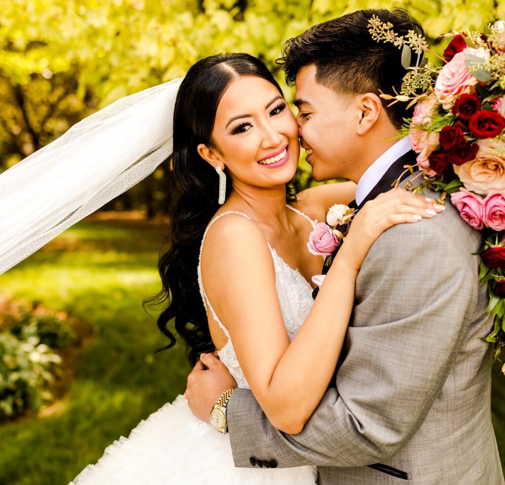 Close-up portrait of Asian bride and groom embracing under floral arch with pink and red roses